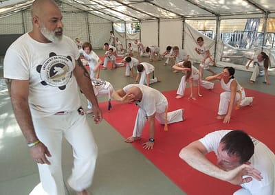 Mestre Nem prowadzi trening Capoeira podczas letniego obozu Capoeira Camangula Poznań. Uczestnicy wykonują wspólnie ćwiczenia techniczne pod okiem doświadczonego mistrza.